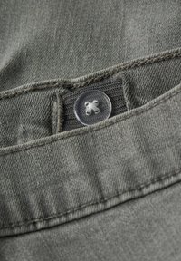 Gråt stof med synlig tekstur og syningsdetaljer. Klar plastknap øverst med fire-huls design.