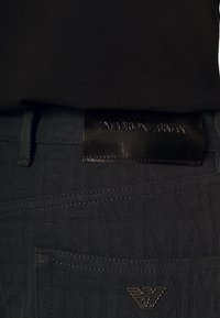 Zbliżenie czarnych spodni Emporio Armani z wytłoczonym wzorem logo, skórzaną metką na pasku oraz metalowym emblematem orła na tylnej kieszeni.