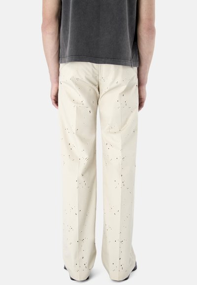 Vista trasera de una persona que lleva pantalones color crema con un patrón de salpicaduras de pintura negra y una camiseta de manga corta gris oscuro.