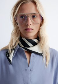 Light blue button-up shirt with a black and white striped scarf around the neck, paired with oversized clear glasses.