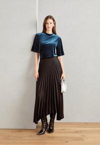 Velvet teal kortemouwen top, zwarte geplooide rok en glanzende knie-hoge laarzen. Houdt een kleine zilveren handtas vast. Neutrale achtergrond.