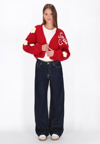 Sarkans adīts piegrieztais kardigans ar ziedu aplikācijām un uzrakstu "Smile" priekšā, kombinēts ar tumši platām biksēm un baltām kedām.