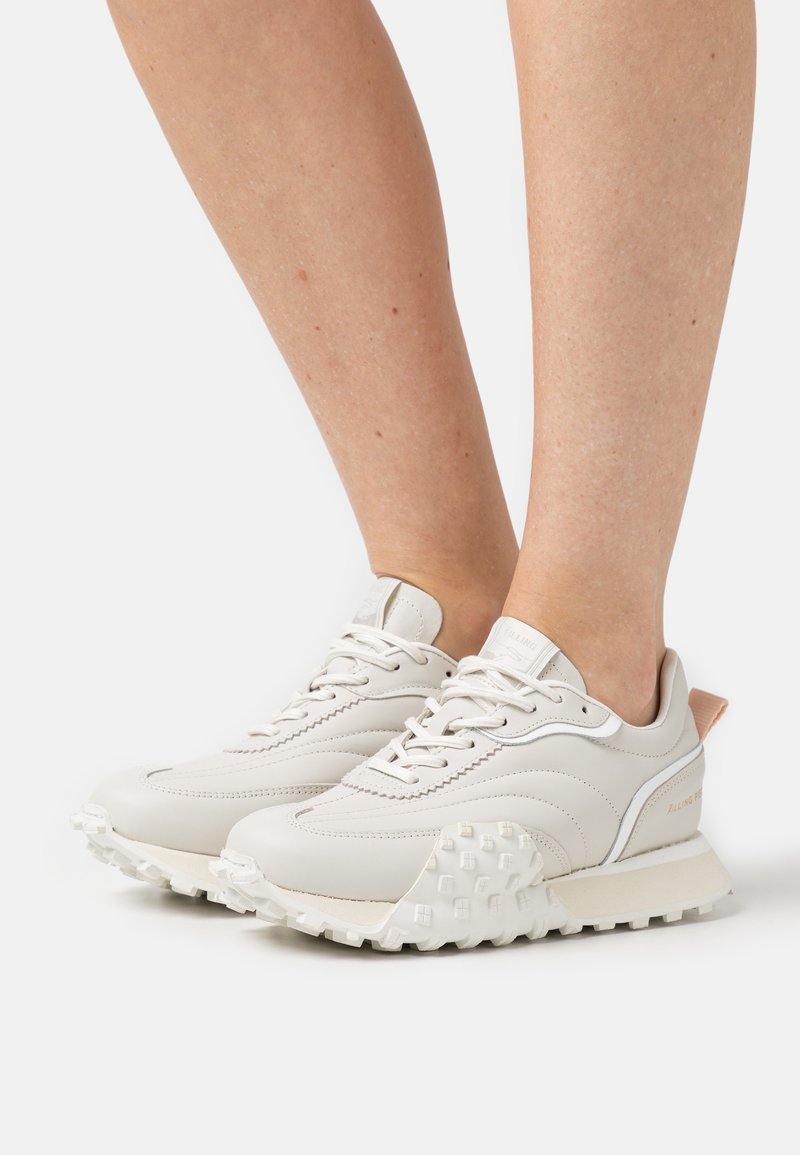 Paire de baskets beiges à lacets avec une semelle texturée portées pieds nus sur un fond blanc uni.