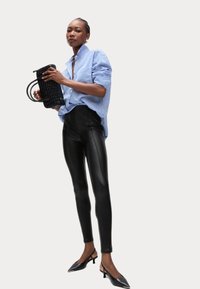 Leggings de piel negro, camisa oversized a rayas azules, bolso negro texturizado y tacones negros puntiagudos, creando un look elegante y moderno.