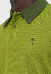 Zöld galléros póló finom bordázott mintával, sötétzöld gallérral és egy kis fekete szív logóval a mellrészen.