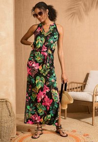 Črna maksi obleka z živahnimi rožnatimi in zelenimi cvetličnimi vzorci, z vrvico v pasu in stranskim razporkom. Manekenka drži tan torbico.