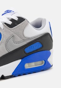 Tenisky Nike Air Max s šedou semišovou a sieťovanou textíliou, čiernymi akcentmi, modrými detailmi a viditeľnou vzduchovou jednotkou v podrážke. Súčasťou sú biele šnúrky.