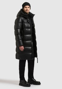 Giacca a vento nera con cappuccio, design imbottito, finitura lucida. Indossata sopra un completo nero con pantaloni testurizzati e stivali neri chunky.