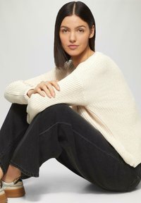 Cremefarbener Pullover aus Strick mit langen Ärmeln, kombiniert mit weiten schwarzen Hosen. Das Model sitzt mit verschränkten Armen und präsentiert eine legere Outfitwahl in entspannter Pose.