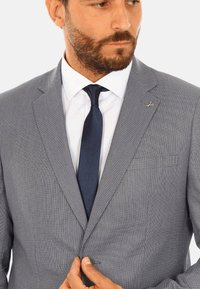 Giacca su misura grigia con un sottile motivo a zig zag, con revers a scoglio e chiusura a un bottone, abbinata a una cravatta scura e una camicia bianca.