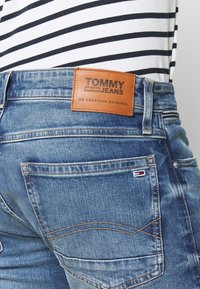 Blå denimjeans med en läderlapp märkt "TOMMY JEANS." Funktioner inkluderar en klassisk design, fem fickor och ton-i-ton sömmar. Randig topp synlig.