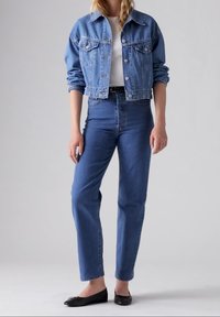 Chaqueta de mezclilla azul claro con cierres de botón y bolsillos en el pecho, combinada con jeans azul claro de cintura alta y bailarinas negras.