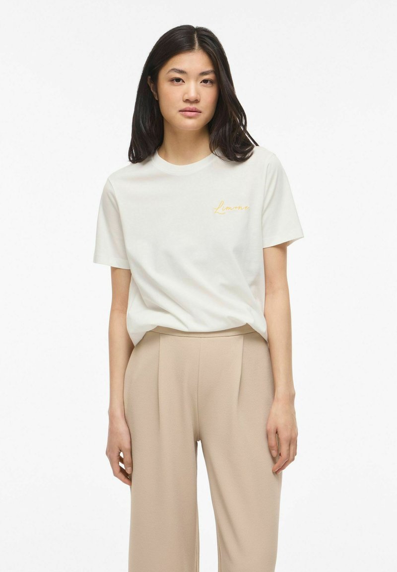 Kvinde med langt mørkt hår iført en hvid T-shirt med gul tekst og beige brede bukser med høj talje, står foran en hvid baggrund.