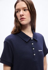 Νεαρή γυναίκα με μακριά καστανά μαλλιά που φοράει μπλε ναυτικό πόλο με λευκά κουμπιά και κεντημένο κείμενο, κοιτάζοντας προς τα αριστερά της.