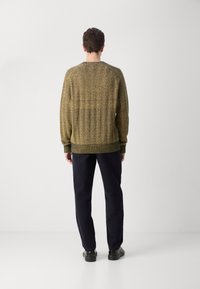 Sweter z teksturowanym splotem w stonowanych żółtych i brązowych odcieniach, z okrągłym dekoltem i ściągaczami na końcach, zestawiony z ciemnymi spodniami i czarnymi butami.