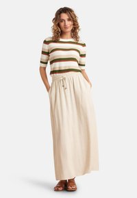 Gebreide gestreepte trui in crème, groen en oranje, gecombineerd met een beige maxi rok met elastische tailleband en zijzakken. Bruine sandalen.