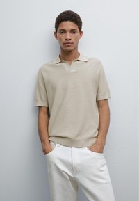 Beige strukturerad polo skjorta med korta ärmar, med en v-ringad krage och ribbad nederkant, i kombination med vita byxor. Enkelt, modernt design.