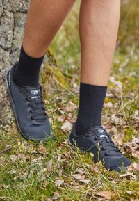 Gambe con calze nere e sneakers nere in piedi su terreno erboso con foglie secche vicino a un tronco di albero.