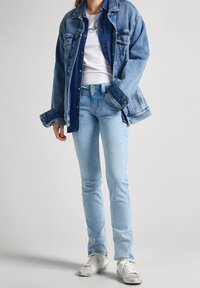 Světle modrá denimová bunda přesažená přes bílou tričko s logem. Slim-fit světle modré džíny a bílé tenisky oblečené dohromady.