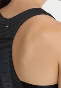 Černý sportovní top bez rukávů s žebrovanou strukturou, širokými ramínky a malým logem poblíž ramene. Zádová část je hladká a obepínající.