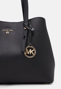 Musta Michael Kors -olkalaukku, jossa on teksturoitu materiaali, kultainen logokiinnike ja irrotettava koriste. Ominaisuuksiin kuuluu pitkät kahvat ja minimalistinen muotoilu.
