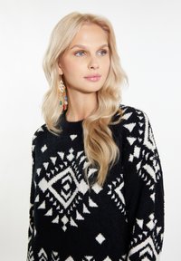 Maglione lavorato a maglia in bianco e nero con motivi geometrici, caratterizzato da una texture morbida. Sono visibili capelli lunghi e biondi e orecchini pendenti colorati.
