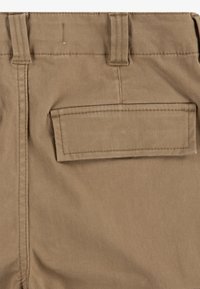 Pantalones de tela tejida beige con un bolsillo trasero estándar, costuras reforzadas y trabillas en la cinturilla. Material de algodón duradero y texturizado.