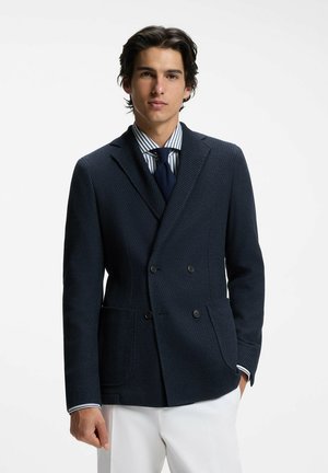 Jeune homme portant un blazer croisé bleu marine, une chemise rayée, une cravate bleu marine et un pantalon blanc, debout sur un fond blanc uni.
