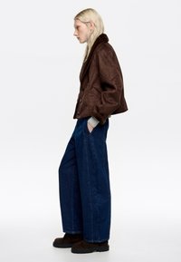 Blondynka w brązowej kurtce z kożuchem, szarym swetrze, szerokich niebieskich dżinsach i ciemnobrązowych butach stoi bokiem z rękami w kieszeniach.