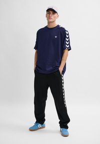 Marineblauw oversized T-shirt met een zijpaneel in wit chevronpatroon, gecombineerd met zwarte broek met witte strepen. Gedragen met blauwe sneakers en een grijze pet.