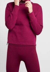 Vínově červená mikina s vysokým límcem, dlouhými rukávy a bočními zipy. Doplněná o ladící legíny vytvářející sladěný sportovní outfit.