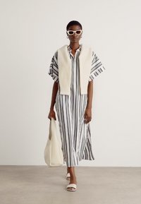 Maxi abito con righe verticali bianche e nere, maniche corte, abbottonatura frontale, abbinato a un avvolgente lavorato a maglia color crema e accessori bianchi.