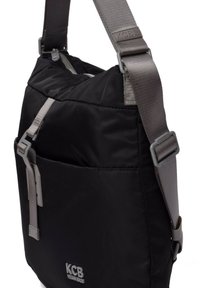 Bandolera negra con un exterior de tela acolchada, correa ajustable gris, bolsillo frontal con cremallera y logo KCB grabado en la parte inferior del frente.
