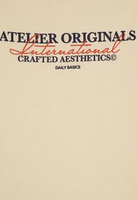 Bežna majica z izrazitim grafičnim besedilom v mornarsko modri in rdeči barvi. Oblikovanje vključuje "ATELIER ORIGINALS" in "CRAFTED AESTHETICS" v izstopajoči pisavi.