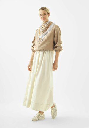 Woman standing, wearing beige long-sleeve top, white ankle-length skirt, patterned scarf, and white sneakers, against a plain white background.