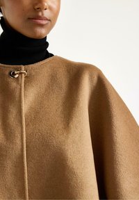Kameluldskappe med en glat tekstur, der har en guldtonet spænde ved halsen og en høj sort turtleneck nedenunder.