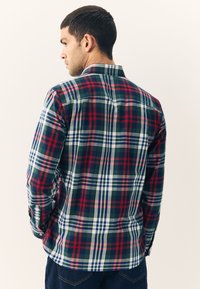 Chemise à carreaux avec un schéma de couleurs vert, rouge, blanc et bleu. Manches longues, col boutonné et ourlet arrondi.