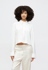Cropped white button-up shirt with a pointed collar, long sleeves, and smooth texture, paired with beige linen trousers.