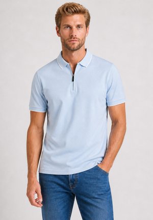 Homme avec une légère barbe et des cheveux coiffés, portant un polo bleu clair à manches courtes zippé et un jean bleu, une main dans la poche.