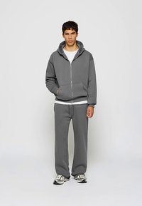 Siva jopa z zadrgo, z velikim kapuco in sprednjimi žepi, v kombinaciji z usklajenimi sivimi trenirkami. Material deluje mehak in teksturiran.