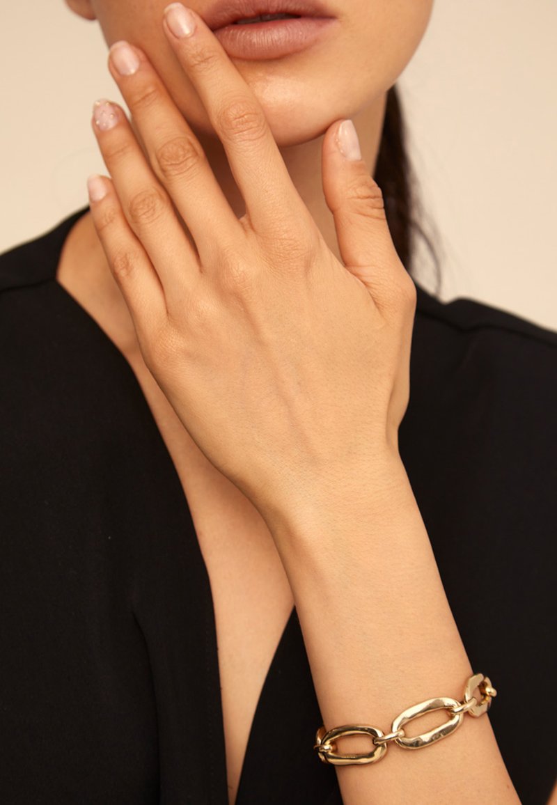 Pulsera de metal dorado con eslabones ovalados interconectados; textura suave con un acabado pulido, llevada en una mano sobre una blusa negra.