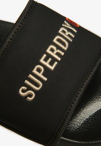 Zwarte slidesandalen met een gevoerde band met geborduurd "SUPERDRY" in goud. Textuurzoel met een subtiel patroon.