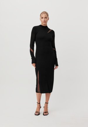Femme portant une robe midi noire ajustée à manches longues avec des découpes transparentes et une fente latérale, associée à des sandales noires à talons hauts et à brides.