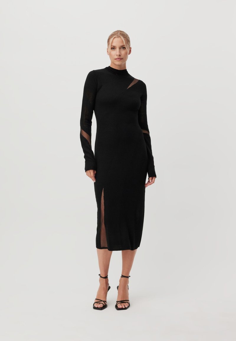 Femme portant une robe midi noire ajustée à manches longues avec des découpes transparentes et une fente latérale, associée à des sandales noires à talons hauts et à brides.