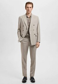 Abito doppio petto color beige chiaro con spalle strutturate, pantaloni slim-fit e una polo abbinata. I mocassini in pelle nera completano il look.