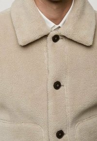 Beige Sherpa-Fleece-Jacke mit großem Kragen, schwarzen Knöpfen und zwei Vordertaschen, getragen über einem weißen Hemd.