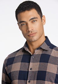 Camisa a cuadros de color azul marino con una combinación de patrones de tartán, que presenta detalles en naranja claro, un cuello abotonado y una textura suave.