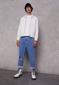 Bílá bomber bunda, bílé tričko, světlé modré džíny s roztržením a bílé šněrovací boty. Uvolněný střih s ohrnutými manžetami na džínách.