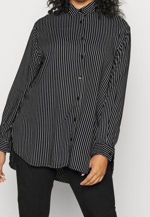 Femme portant une chemise noire ample à boutons avec des rayures blanches verticales et un pantalon noir sur un fond clair uni.
