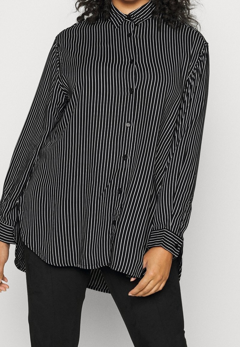 Femme portant une chemise noire ample à boutons avec des rayures blanches verticales et un pantalon noir sur un fond clair uni.